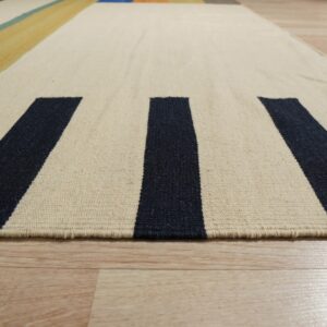 Cream and navy striped flatweave rug texture detail on light wood flooring, with distant yellow and teal blocks.