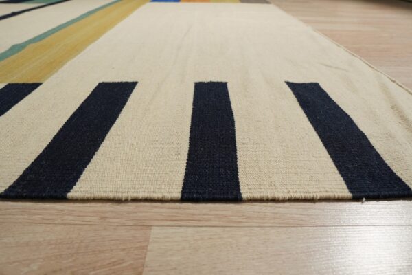 Cream and navy striped flatweave rug texture detail on light wood flooring, with distant yellow and teal blocks.