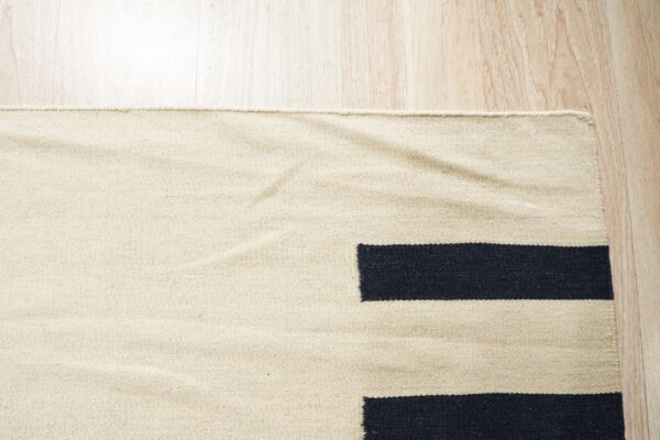 Corner of a flatwoven cream and dark geometric rug resting on light wood flooring.