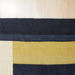 Flatweave rug with blocks of navy blue, gold, and cream on light wood plank flooring.