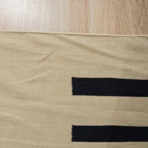 A flat-weave beige rug with large geometric black bars rests on light wood flooring.