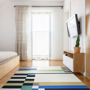 Low-pile geometric rug with blue, teal, orange, and navy blocks covers wood floor near a window.