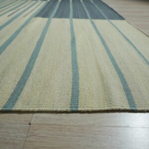 Low-pile rug in cream with teal stripes and a dark blue color block on light wood flooring.