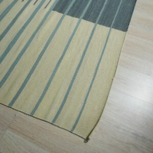 Striped flatweave rug in pale yellow and teal with a dark gray section resting on light blonde plank flooring.