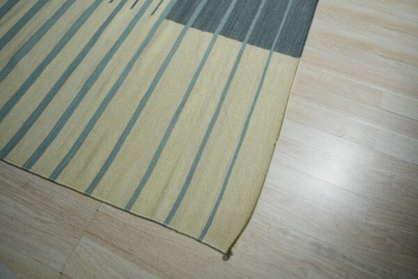 Striped flatweave rug in pale yellow and teal with a dark gray section resting on light blonde plank flooring.