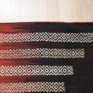Dark brown and terracotta rug with white geometric diamond stripes rests on light wood floor.