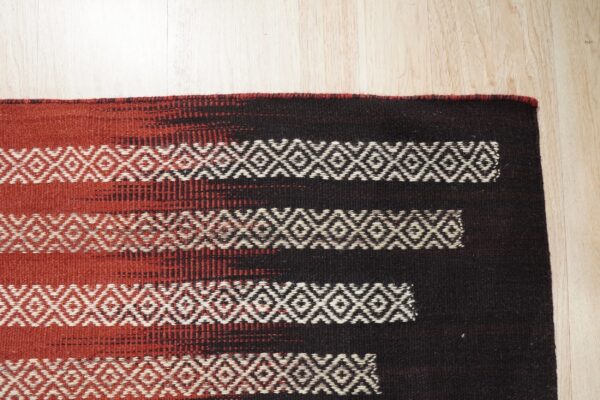 Dark brown and terracotta rug with white geometric diamond stripes rests on light wood floor.