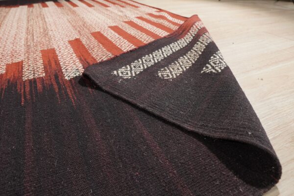Detail shot of a flatweave rug showing radiating rust and white patterns transitioning to dark brown on light wood flooring.