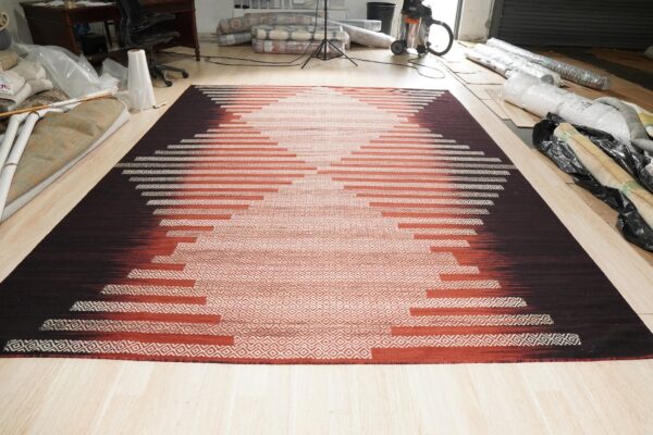 Flatweave rug with a stepped geometric pattern transitioning from dark brown to cream on light wood flooring.