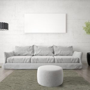 A muted green lattice rug anchors a grey sofa and ottoman against a white textured wall and wood floor.