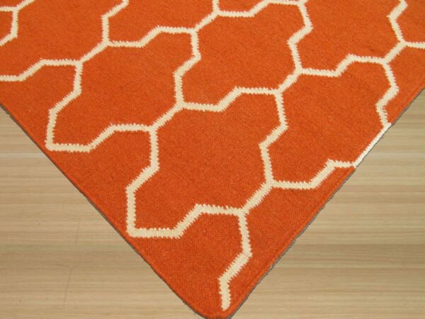 Orange low-pile rug displaying a cream interlocking geometric pattern on light blonde wood flooring.