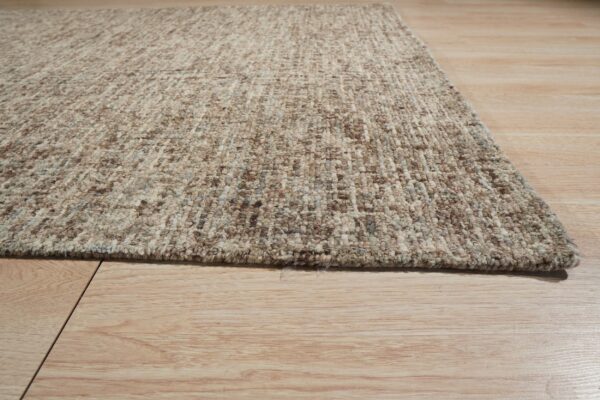 A close-up of the speckled, textured beige and brown rug edge on light wood flooring.