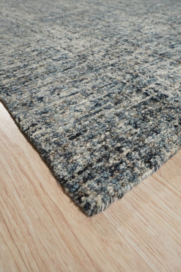 A corner of a chunky textured rug showing mixed blue, gray, and cream colors on light hardwood.