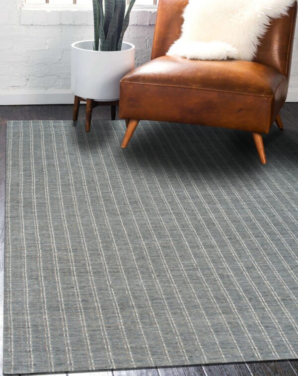 Low pile blue rug with thin vertical stripes next to a brown leather armchair and white brick wall.