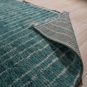 Close-up shows a high-pile teal rug with white abstract lines curled back on blonde wood floor.