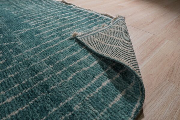 Close-up shows a high-pile teal rug with white abstract lines curled back on blonde wood floor.