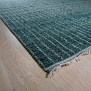 High-pile teal rug with an off-white geometric grid and visible fringe rests on light wood plank flooring.