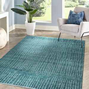 A teal rug with an abstract white striped pattern sits on herringbone wood flooring next to a gray chair and a large window.