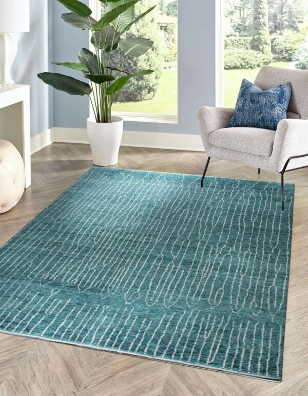 A teal rug with an abstract white striped pattern sits on herringbone wood flooring next to a gray chair and a large window.