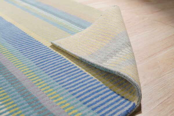 Blue, yellow, and teal low-pile rug with broken stripes is curled back on light wood flooring.