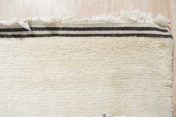 High-pile cream rug with black striped flat-woven edge and white fringe on light wood floor.
