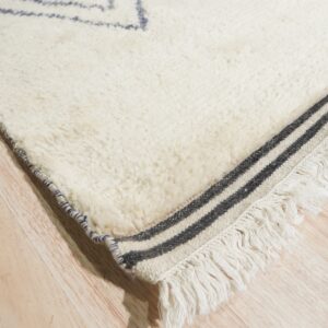 Cream shaggy rug corner showing blue diamond motifs, striped woven fringe, and wood planks.
