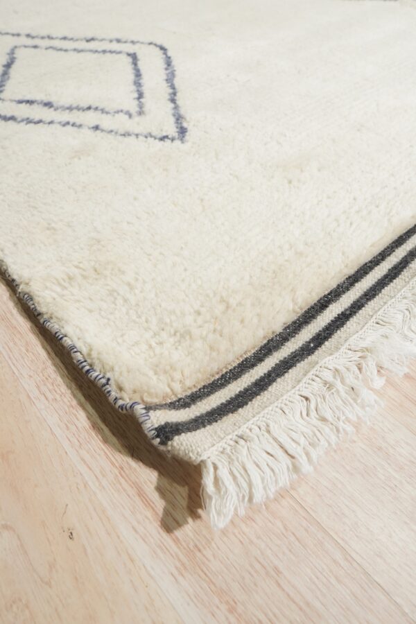 Cream shaggy rug corner showing blue diamond motifs, striped woven fringe, and wood planks.