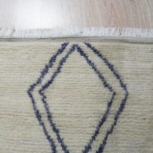 Textured off-white rug showing a blue geometric pattern and fringe over light wood planks.