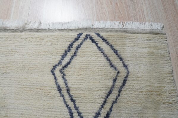 Textured off-white rug showing a blue geometric pattern and fringe over light wood planks.