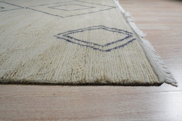 Textured off-white rug with dark blue geometric diamond motifs and fringe on pale wood floors.