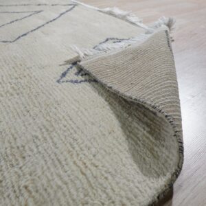 Shaggy cream rug with thin dark blue patterns, corner lifted to expose the woven backing on light flooring.