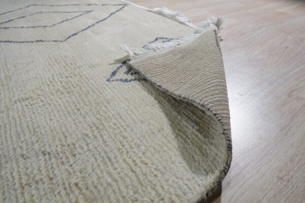 Shaggy cream rug with thin dark blue patterns, corner lifted to expose the woven backing on light flooring.