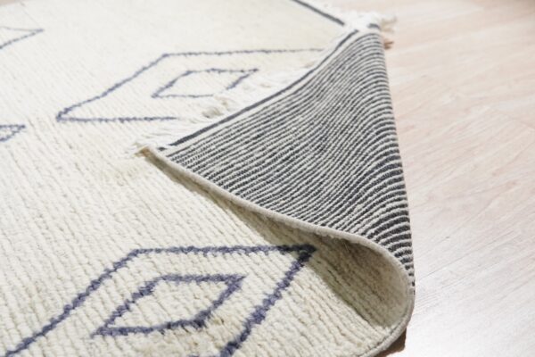 High-pile cream rug with blue diamonds is folded back, showing its striped backing on light wood.