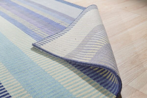 Rug with multicolor blue, lavender, and cream stripes is partially rolled on light hardwood flooring.