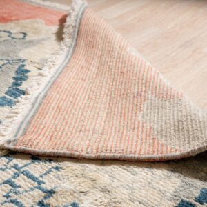 High-pile rug with blue and cream pattern curled on wood floor, exposing the striped peach backing.
