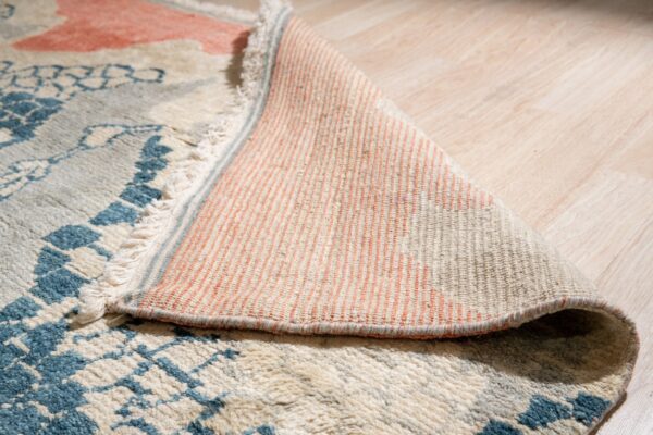 High-pile rug with blue and cream pattern curled on wood floor, exposing the striped peach backing.