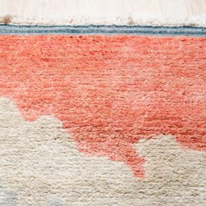 Close-up of a high-pile rug with abstract pink-orange and beige shapes, fringe, and blue binding on light wood flooring.