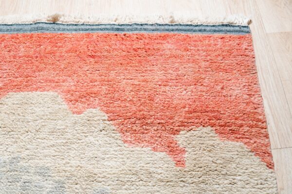 Close-up of a high-pile rug with abstract pink-orange and beige shapes, fringe, and blue binding on light wood flooring.