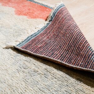 Shaggy off-white rug with coral and blue accents, revealing its striped woven backing on wood flooring.