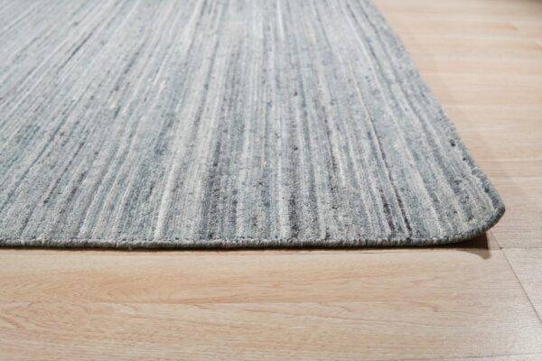 A striated blue and gray low-pile rug rests with a rounded edge on light blonde wood plank flooring.