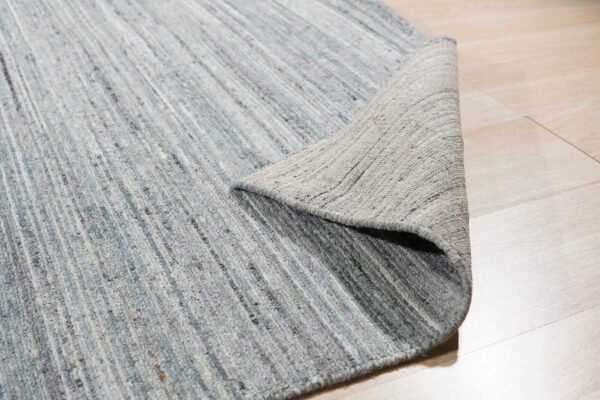 Blue and gray low-pile rug with vertical striping, partially curled back on light wood flooring.