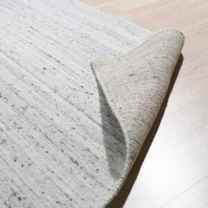 Textured light gray rug with dark flecks partially rolled, showing the backing on light wood plank flooring.