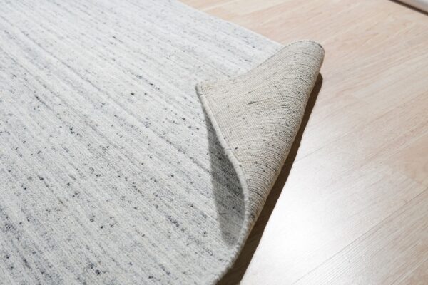 Textured light gray rug with dark flecks partially rolled, showing the backing on light wood plank flooring.