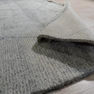 High-pile light gray rug with faint geometric lines, rolled up to reveal the woven tan backing.