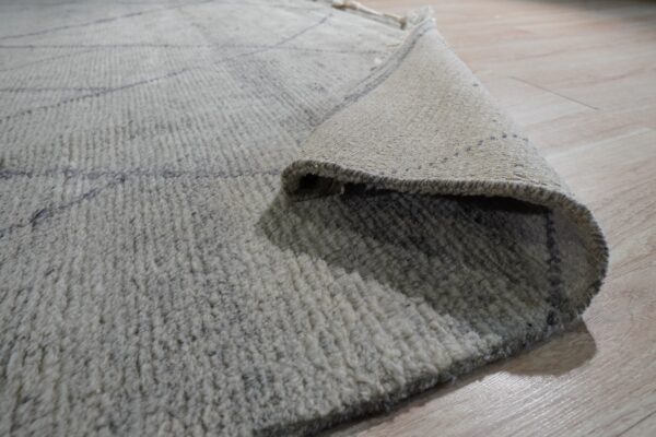 High-pile light gray rug with faint geometric lines, rolled up to reveal the woven tan backing.