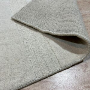 Close-up view of a light beige rug with visible pile height variation, folded over revealing the woven backing on wood flooring.