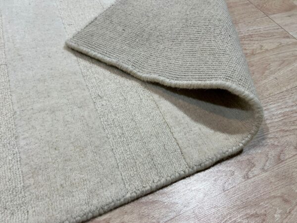 Close-up view of a light beige rug with visible pile height variation, folded over revealing the woven backing on wood flooring.