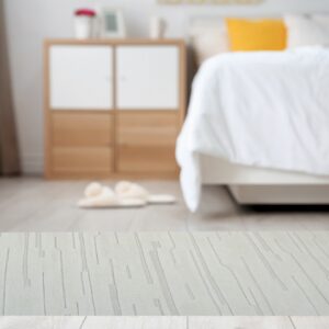 A light beige rug with an abstract carved linear pattern rests on light wood flooring in a blurred bedroom.