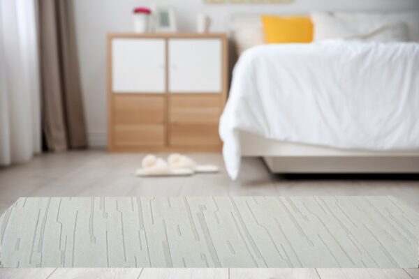 A light beige rug with an abstract carved linear pattern rests on light wood flooring in a blurred bedroom.