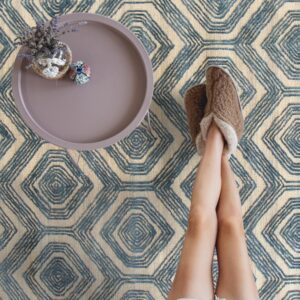 A textured blue and cream rug with a geometric hexagonal pattern underlies a person's legs and slippers.